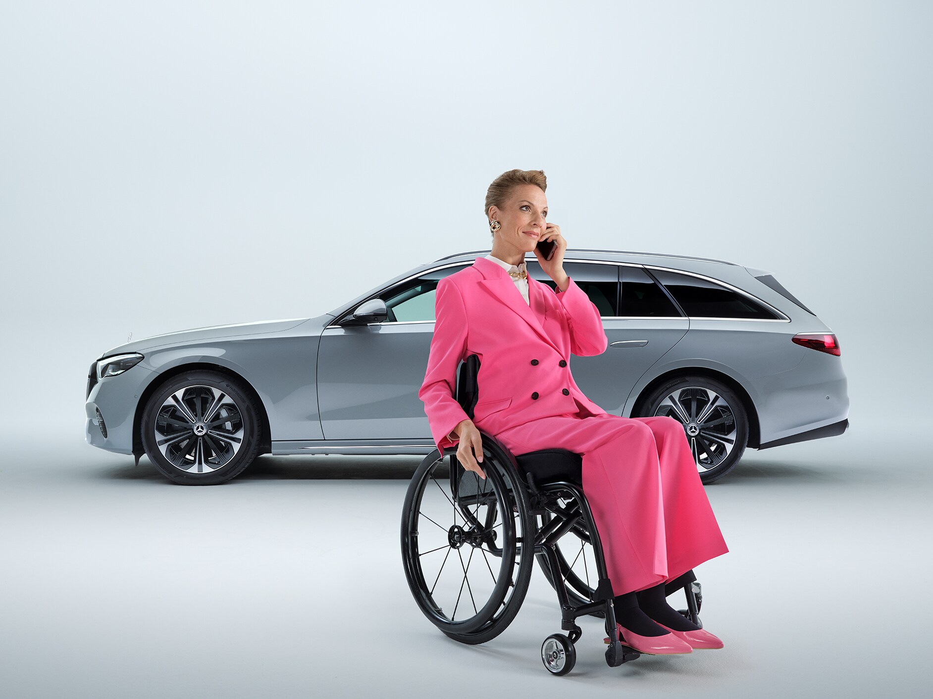 Vue latérale d'un break Mercedes-Benz Classe E transformé en voiture pour personnes à mobilité réduite, avec au premier plan une femme en fauteuil roulant tenant un smartphone.