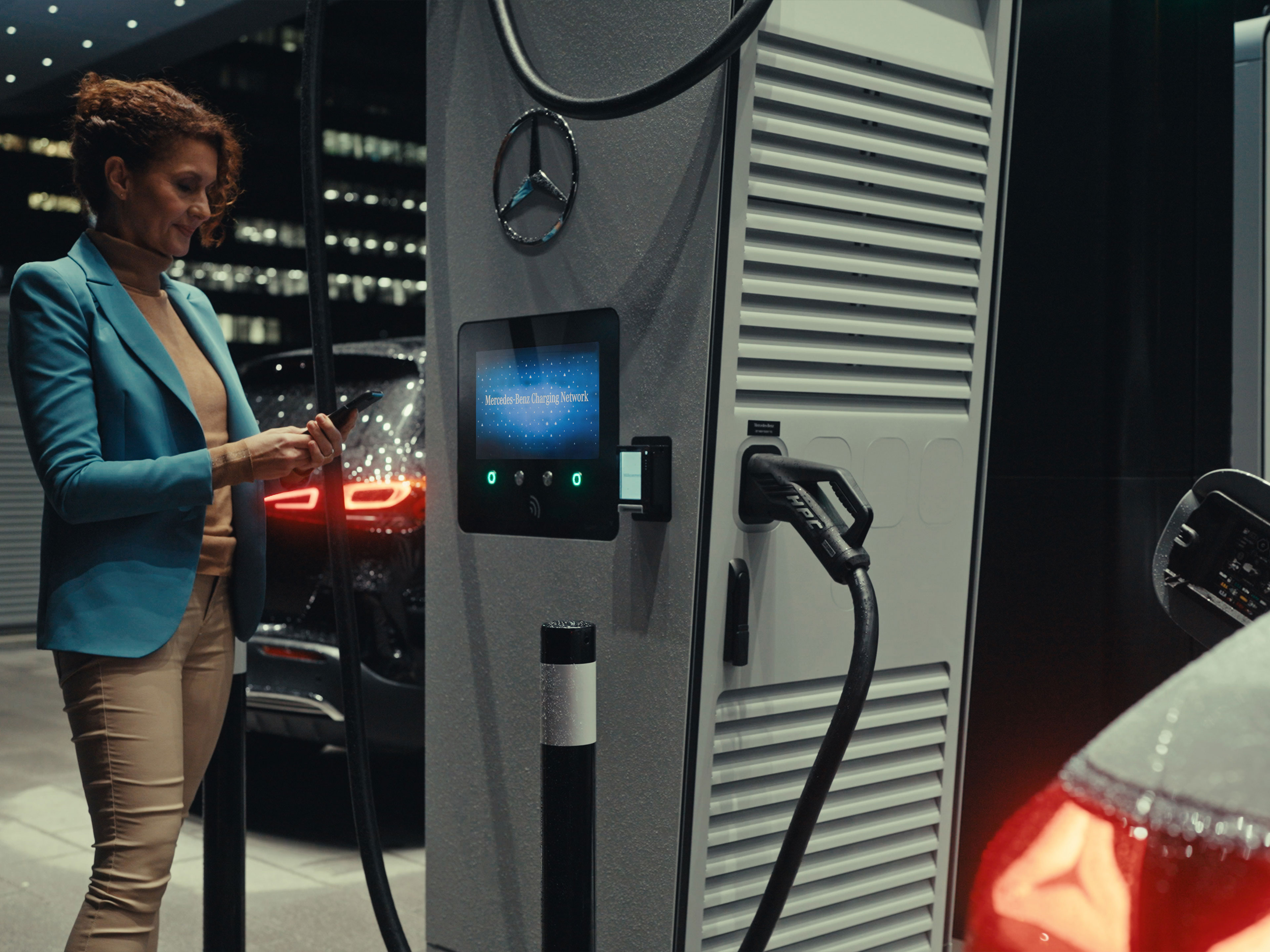 Mercedes-Benz App | Laden unterwegs | Mercedes-Benz Eine Frau, die ein Smartphone in der Hand hält und neben einer Ladestation steht.