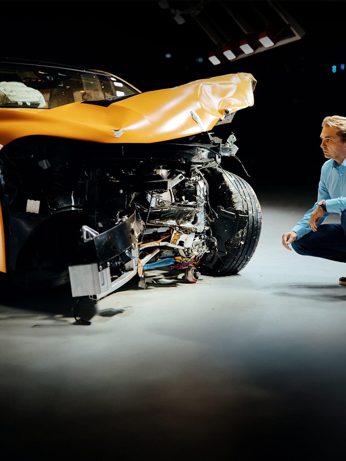 Un expert en essais de collision examine une voiture Mercedes-Benz accidentée à l'avant en s'accroupissant à côté d'elle.