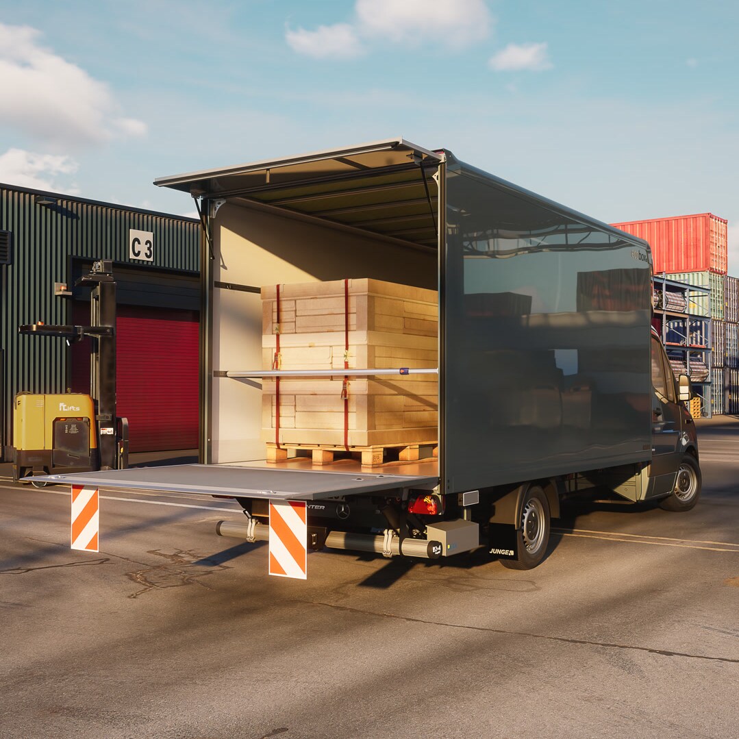 Une Mercedes-Benz Sprinter équipée d'une caisse pour vrac sec de la marque Junge lors d'une opération de chargement dans un centre logistique – une solution de transport efficace pour le fret général et la manutention de marchandises. La porte arrière ouverte laisse apparaître des panneaux de bois palettisés dans le compartiment de chargement pendant le chargement ou le déchargement.