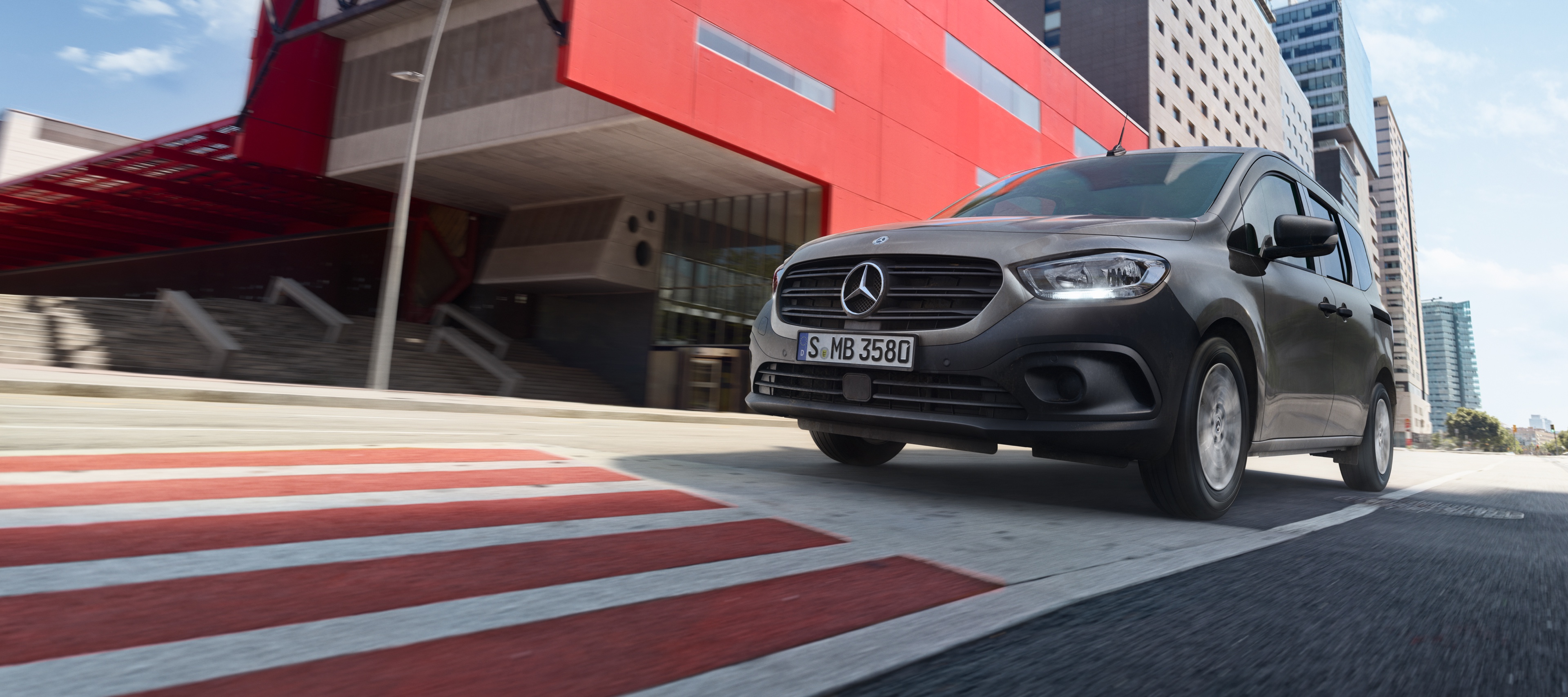 Citan Tourer | Veicoli Commerciali Mercedes-Benz Il Citan Tourer, un furgone per il trasporto passeggeri Mercedes-Benz, passa attraverso un quartiere urbano moderno.