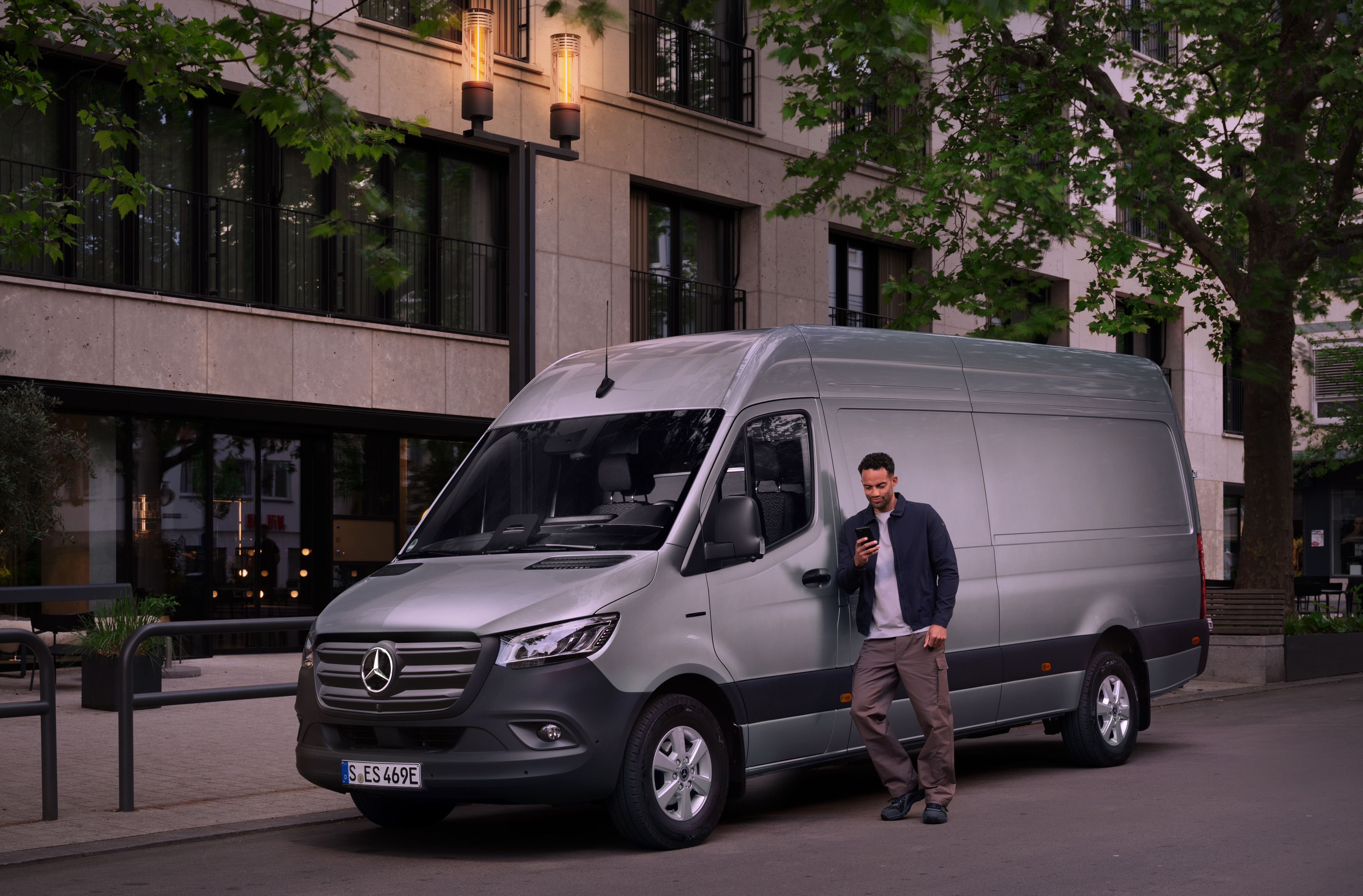 Mercedes-Benz | Assistance pour utilitaires légers | Centre d'assistance à la clientèle Un homme s'appuie sur un utilitaire léger Mercedes-Benz Sprinter argenté et utilise son smartphone - assistance rapide du service clientèle de Mercedes Benz.