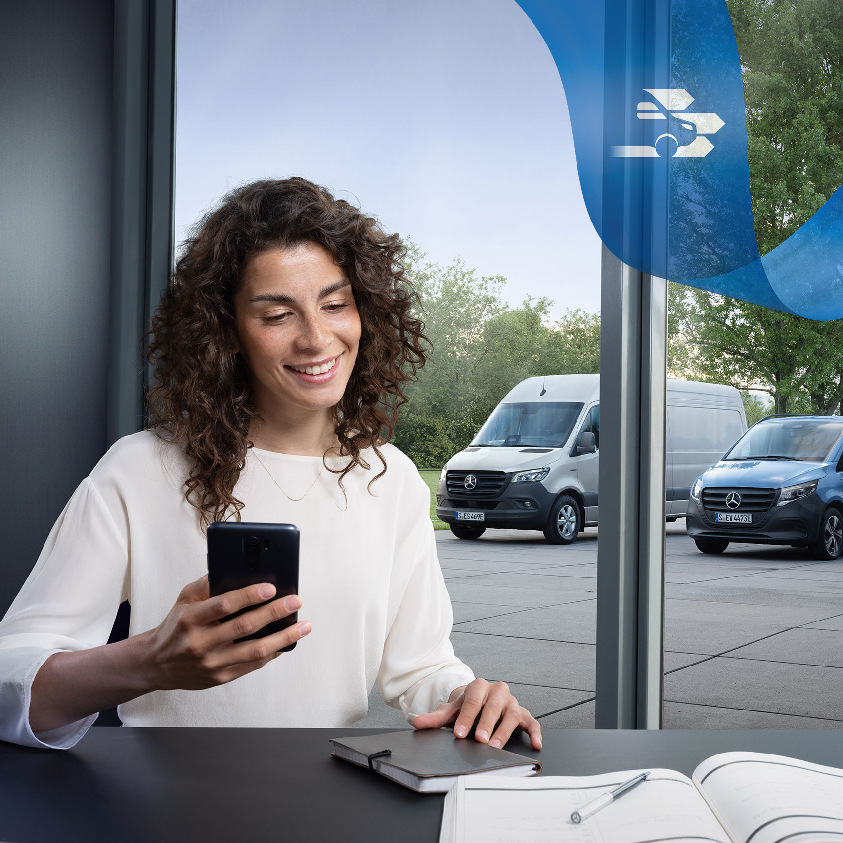 Mercedes-Benz | Assistenza veicoli commerciali | Centro assistenza clienti Una donna seduta alla scrivania con uno smartphone e una serie di veicoli commerciali Mercedes-Benz sullo sfondo.