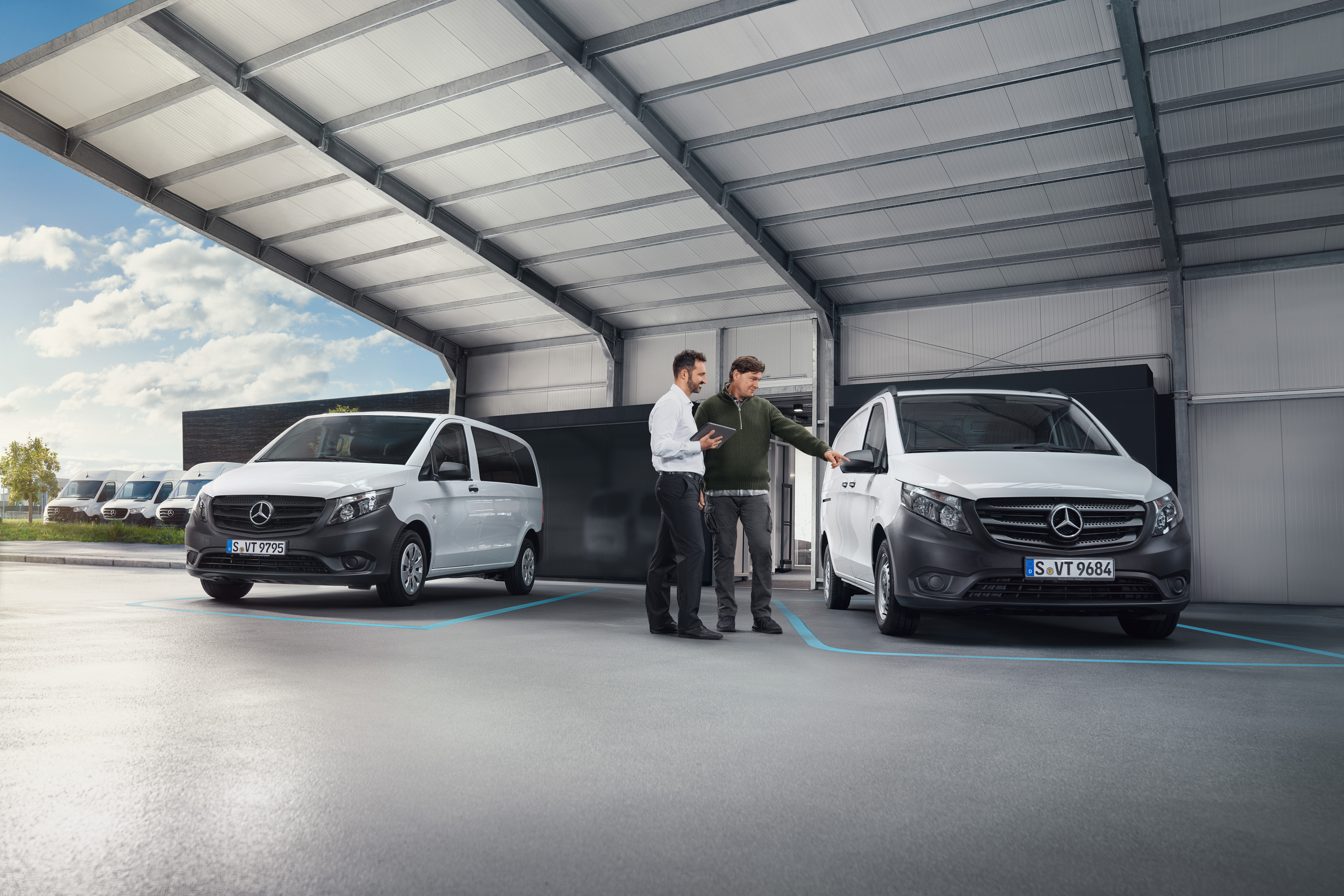 Mercedes-Benz | Convenience | Accettazione digitale dei clienti Due persone mentre parlano in piedi in un ufficio davanti a due veicoli commerciali Mercedes-Benz bianchi.