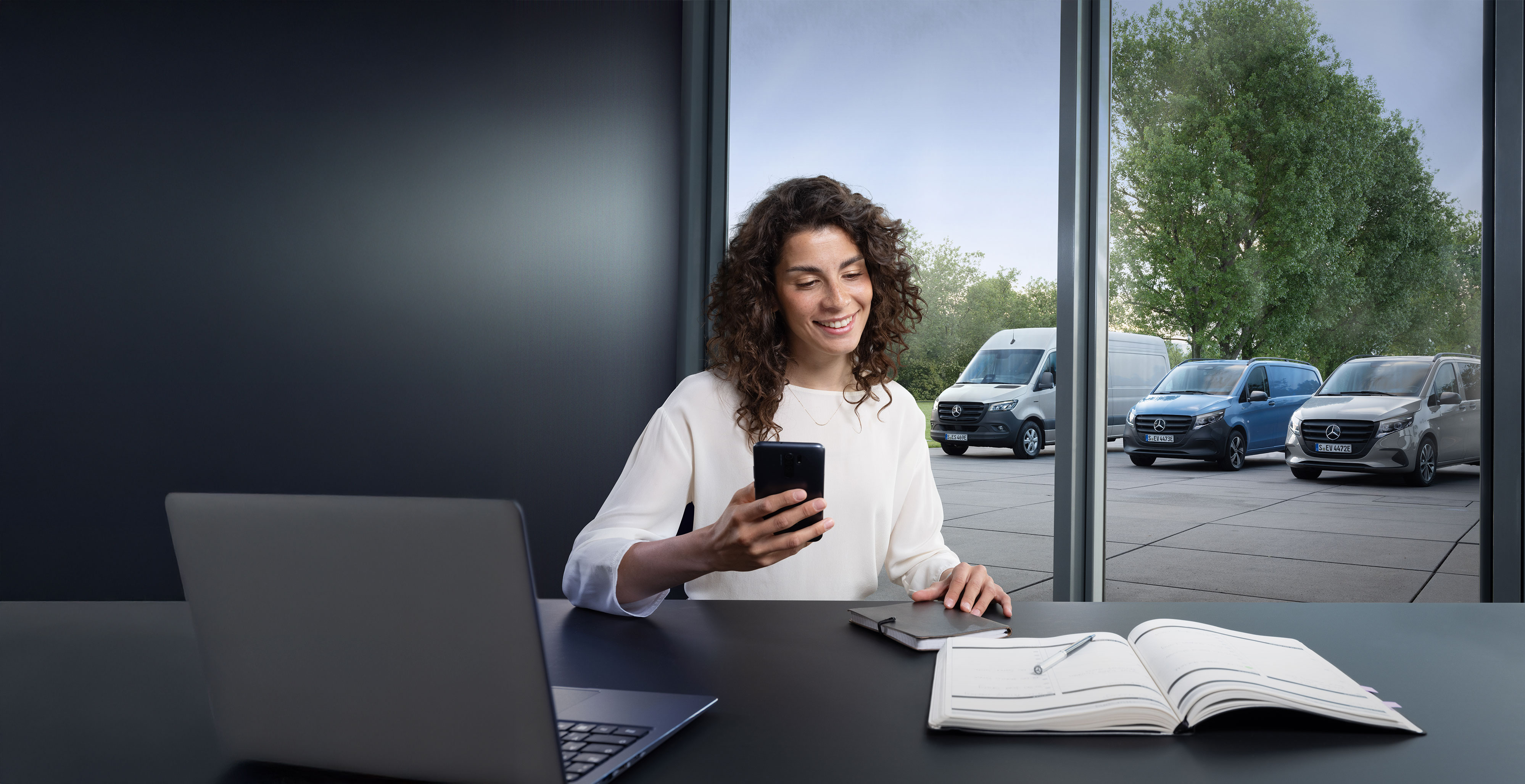 Mercedes Benz | Convenience | Prenotazione appuntamenti online Una donna mentre prenota un servizio assistenza tramite smartphone; sullo sfondo una serie di veicoli commerciali Mercedes-Benz.