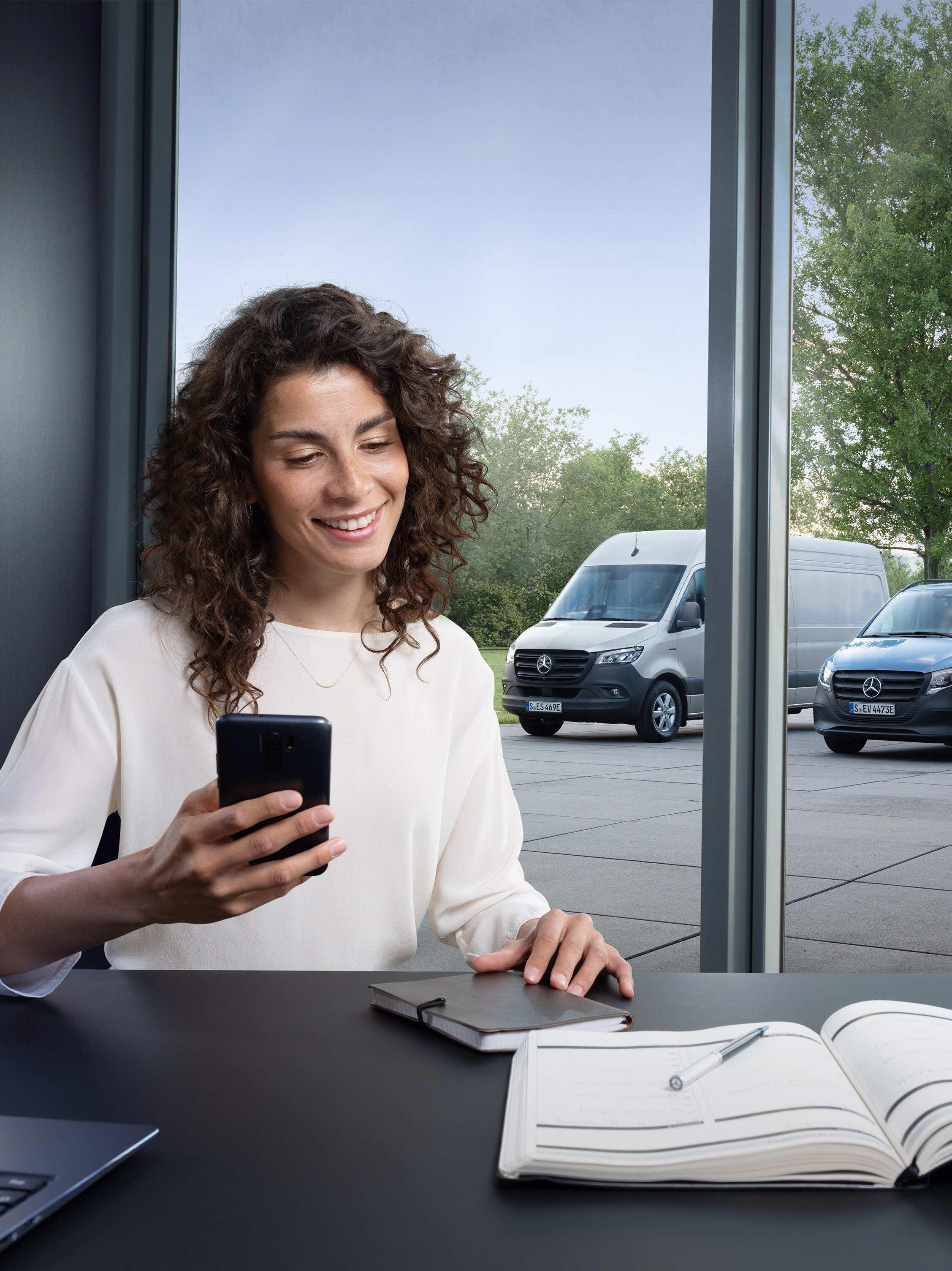 Femme avec un smartphone devant une fenêtre avec trois utilitaires légers Mercedes-Benz à l'extérieur.