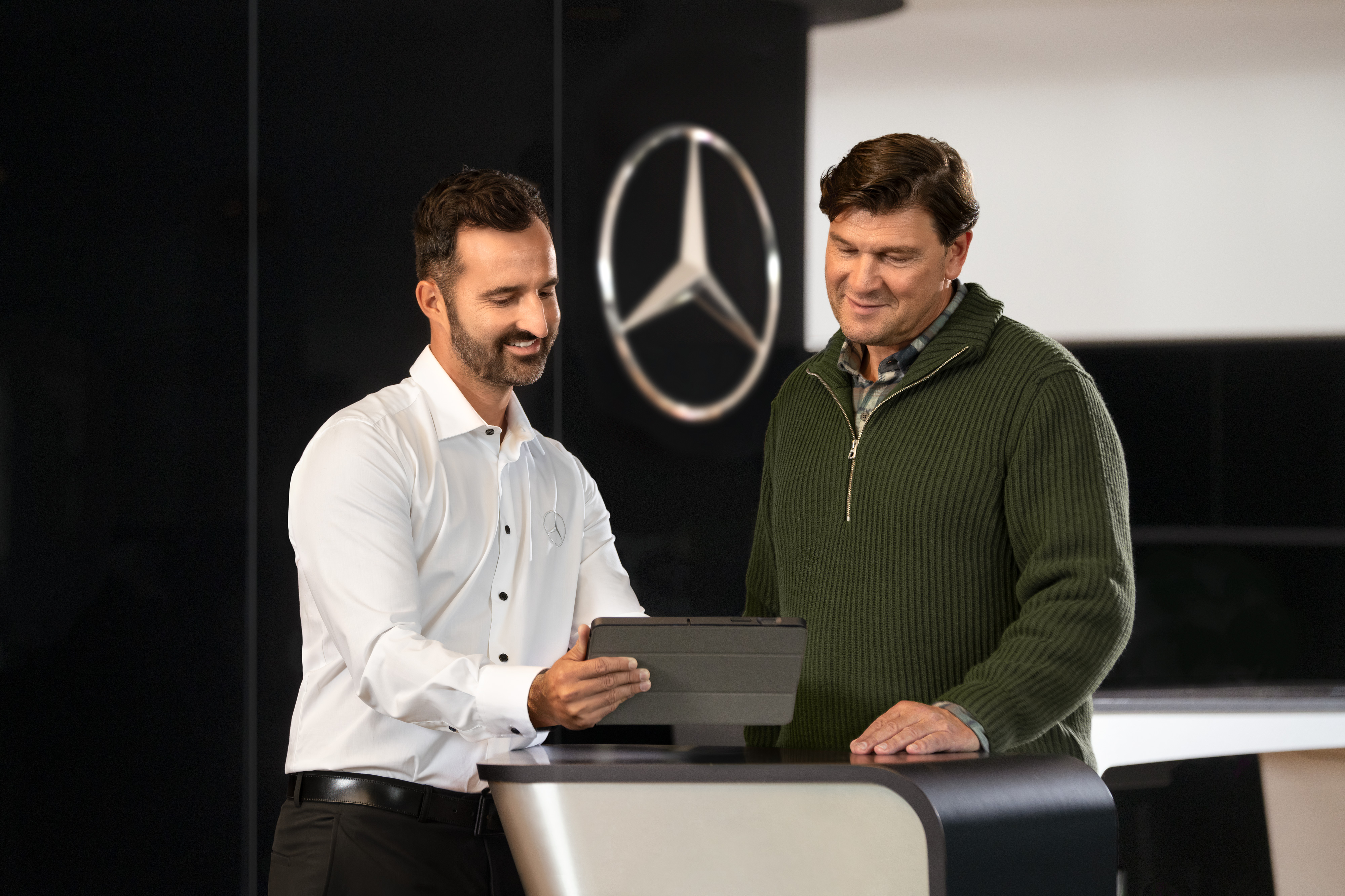 Mercedes-Benz | Services Overview Due uomini parlano a un bancone mentre guardano un tablet; davanti a loro la Stella Mercedes-Benz.