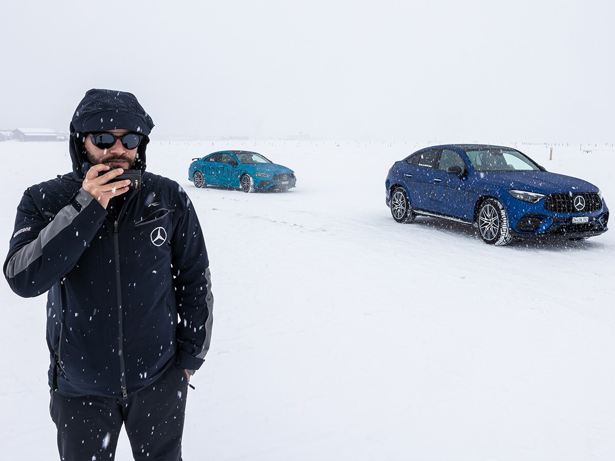 Domenico Solombrino, veste Mercedes-Benz noire et lunettes de soleil, se tient sur une piste d’essai enneigée. Deux modèles Mercedes-Benz bleus sont visibles en arrière-plan.