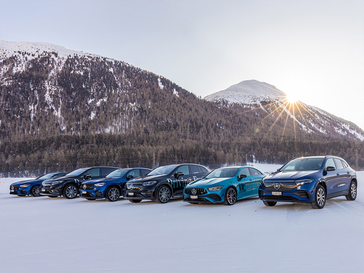 Die Mercedes-Benz Winterflotte Eine Reihe von Mercedes-Benz Fahrzeugen steht in Formation auf einer verschneiten Ebene vor verschneiten Wäldern und tiefstehender Wintersonne.