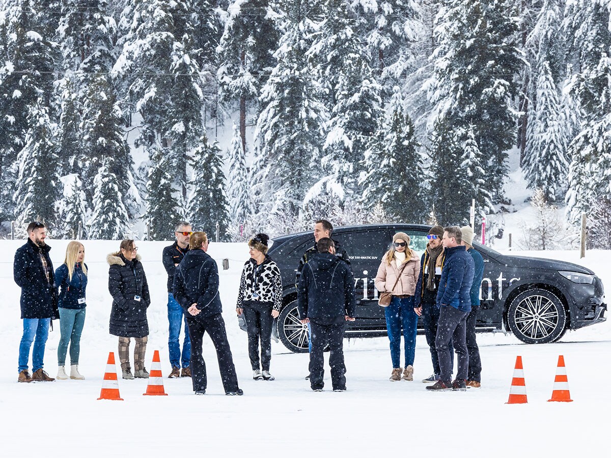 Plusieurs participants à la Winter Experience, vêtus de tenues d’hiver, se tiennent à côté d’un SUV Mercedes-Benz noir, au milieu d’une forêt recouverte de neige.
