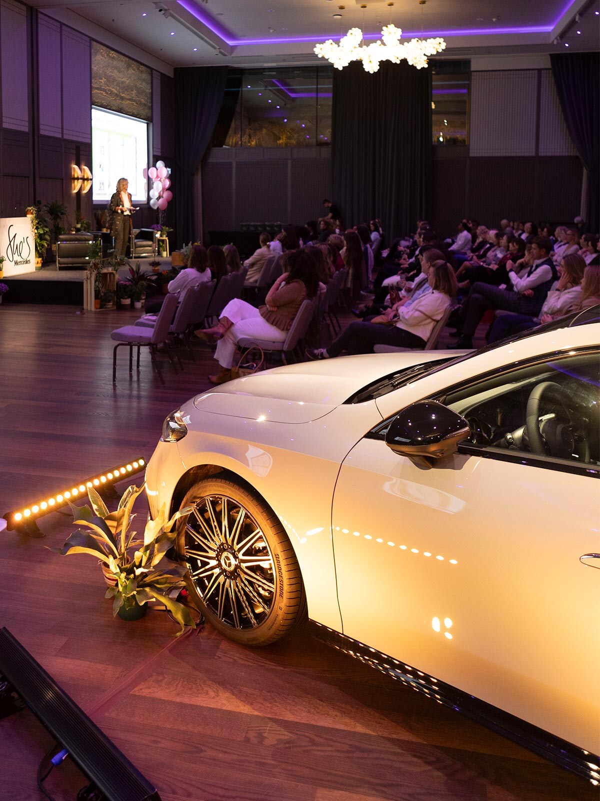 Salle de l’événement avec invités, conférencière et Mercedes blanche.