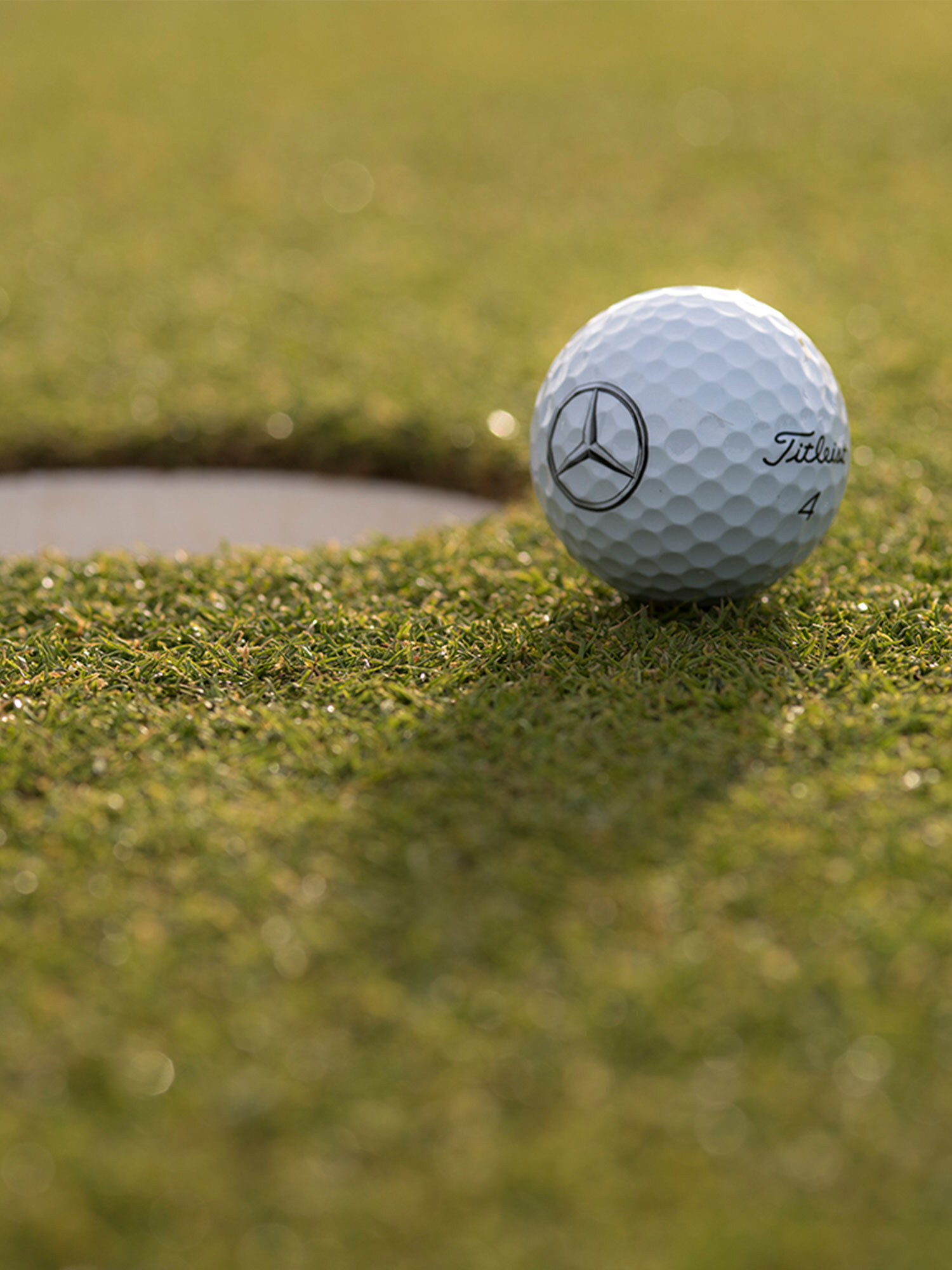 Una palla da golf con il logo Mercedes-Benz si trova appena prima della buca su un prato ben curato.