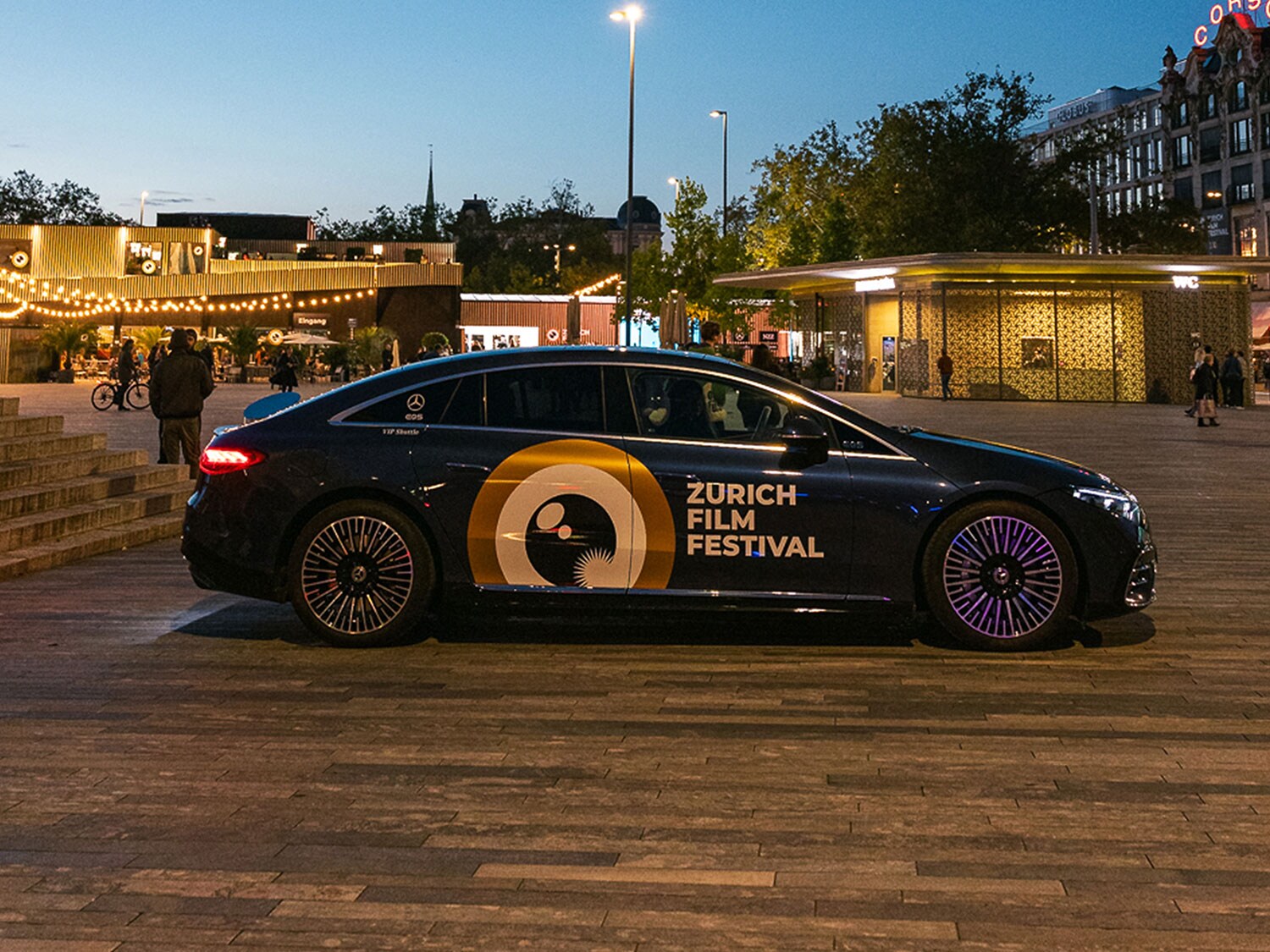Ein Mercedes EQS mit Zurich Film Festival Branding steht bei Abenddämmerung auf dem Sechseläutenplatz, umgeben von stimmungsvoll beleuchteten Pavillons und Passanten.