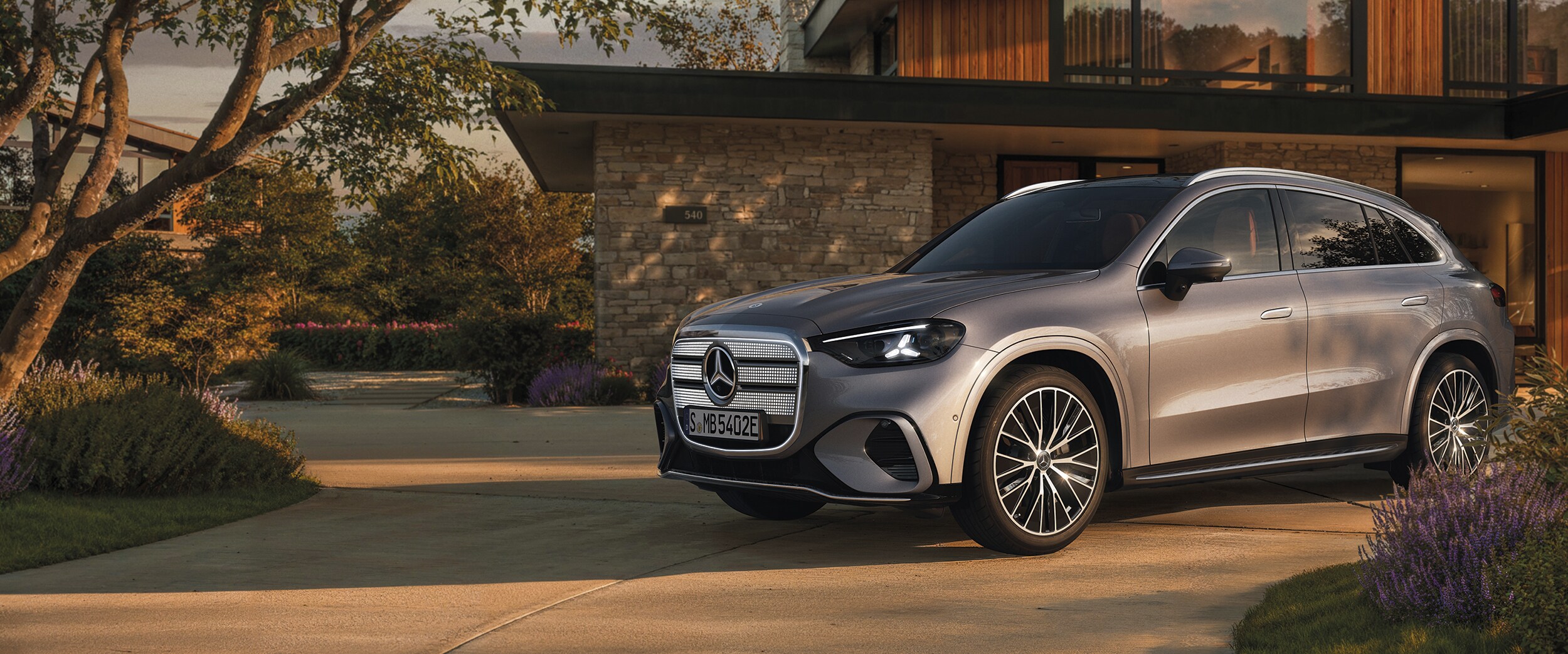Le GLC entièrement électrique de Mercedes-Benz se tient devant une maison moderne, au coucher du soleil, au bout d’une allée courbe.