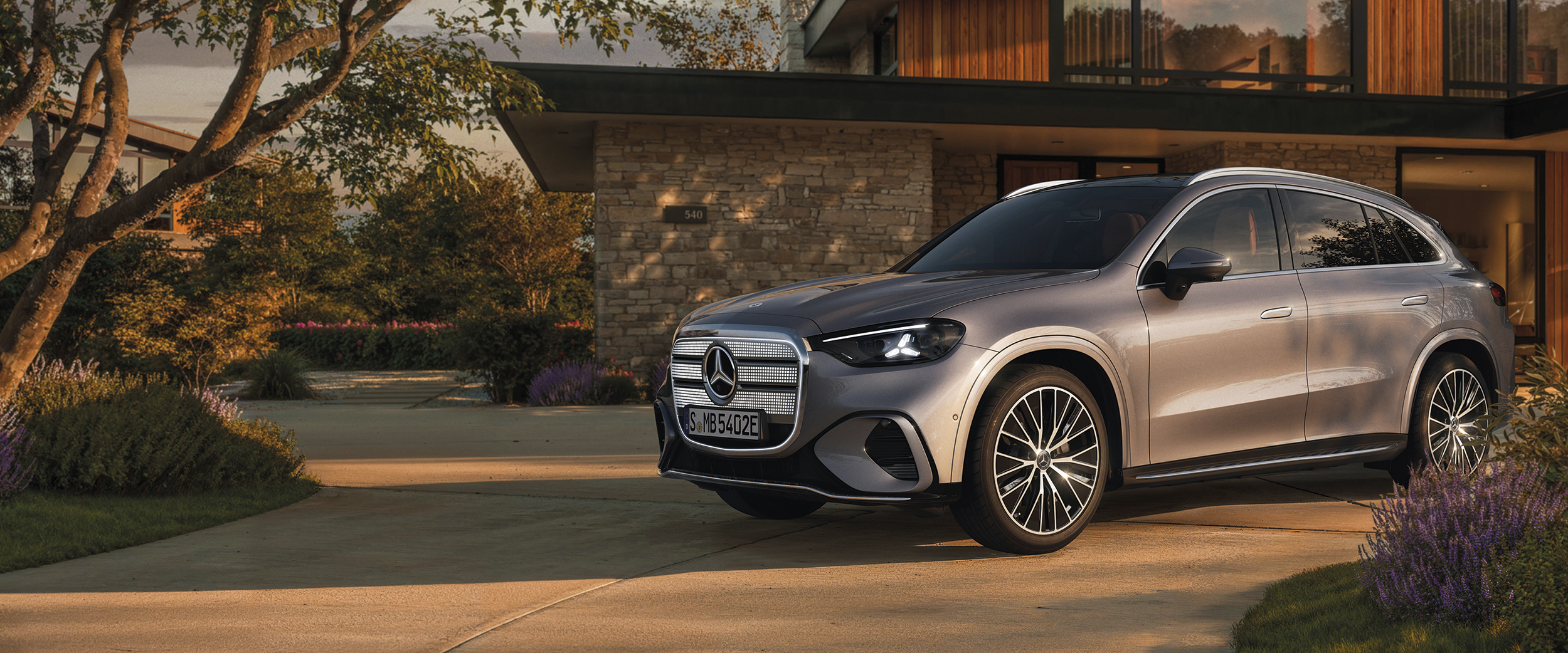 Le nouveau GLC entièrement électrique Le GLC entièrement électrique de Mercedes-Benz se tient devant une maison moderne, au coucher du soleil, au bout d’une allée courbe.