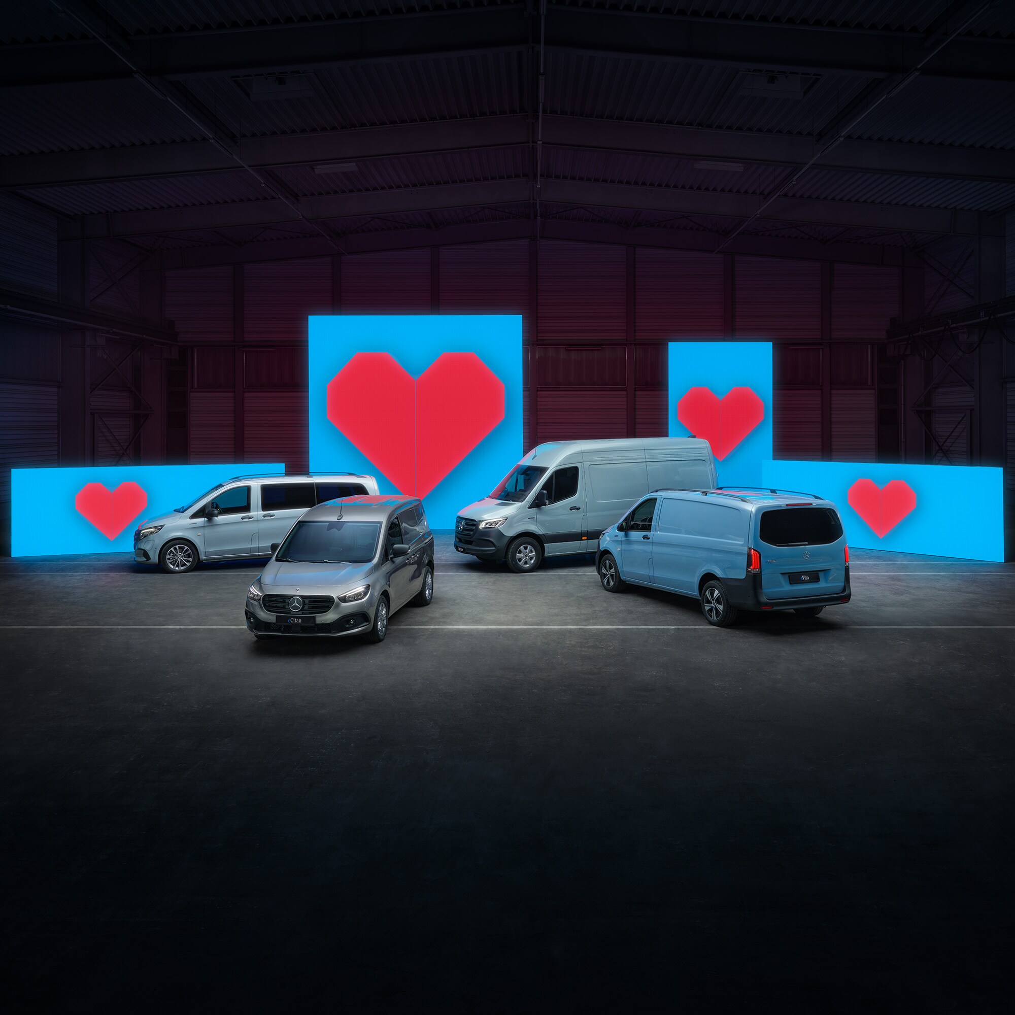 Verschiedene Van-Elektromodelle stehen vor blauen Leuchtwänden mit je einem roten Herz.