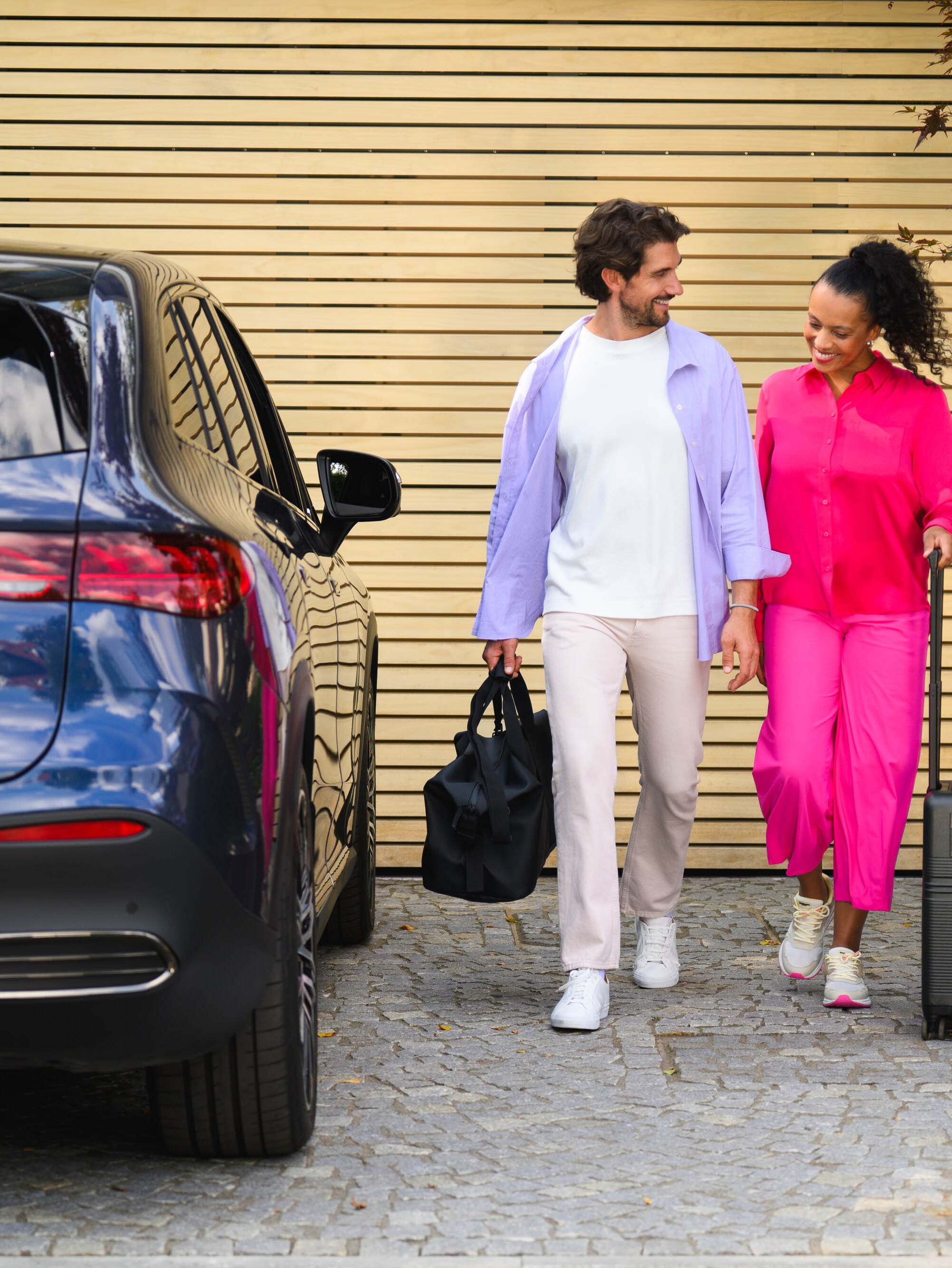 Zwei Personen gehen auf einen Mercedes-Benz zu, eine mit einem Koffer und die andere mit einer Sporttasche.