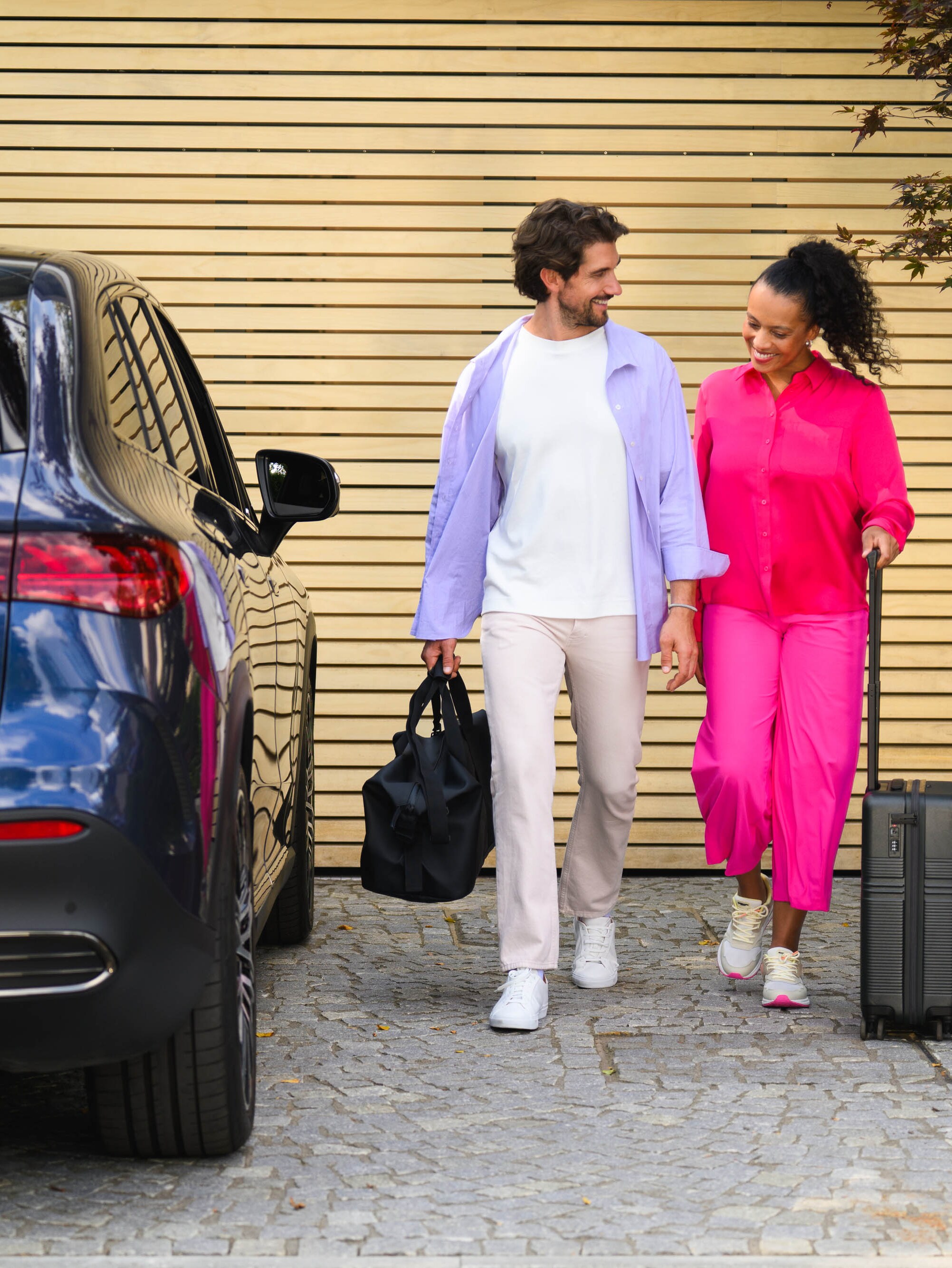 Zwei Personen gehen auf einen Mercedes-Benz zu, eine mit einem Koffer und die andere mit einer Sporttasche.