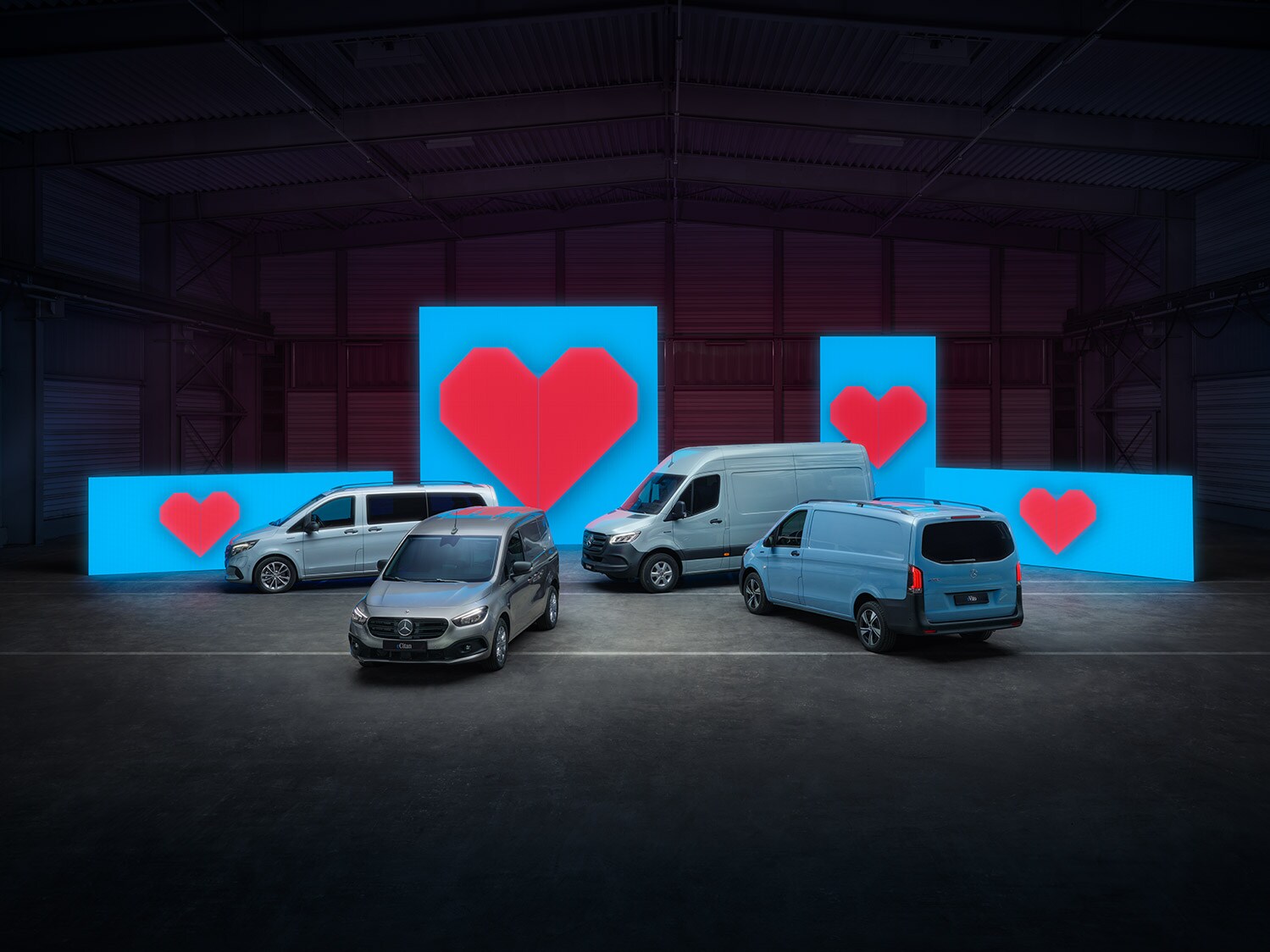 Vari modelli di van elettrici si trovano davanti a pareti illuminate di blu, ognuno con un cuore rosso.